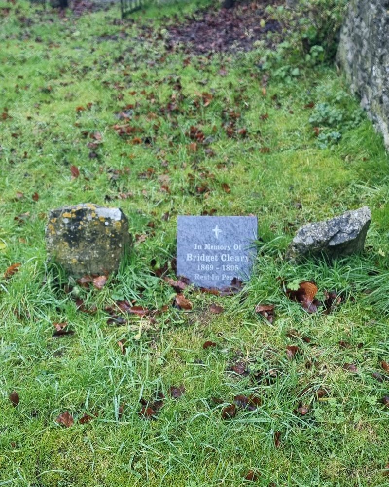 A small slate memorial plaque reading In Memory Of Bridget Cleary set in the grass between two rough stones in a country graveyard