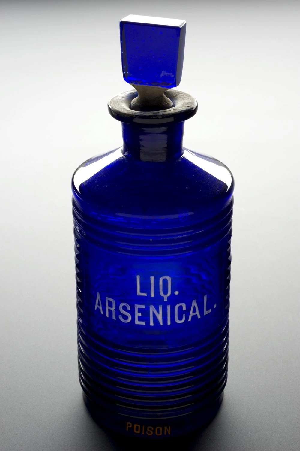 A ridged cobalt blue glass pharmacy bottle with a square stopper, labeled LIQ. ARSENICAL and POISON