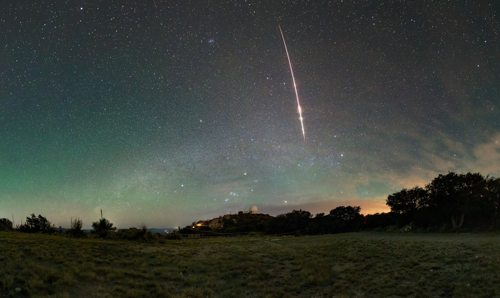 A bright fireball streaks diagonally across a blue-green predawn sky above the domes of Kitt Peak National Observatory in Arizona