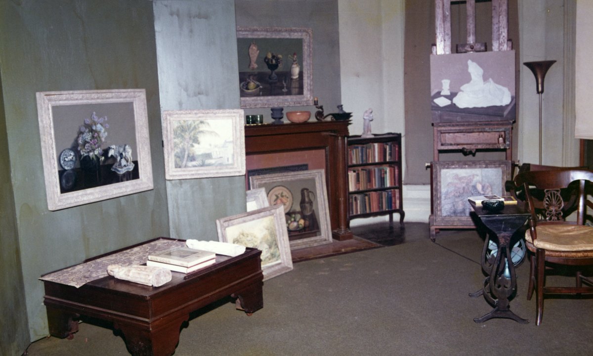 A room with framed still-life paintings on the walls and floor, an easel, a fireplace, a bookcase and a cane-seated chair