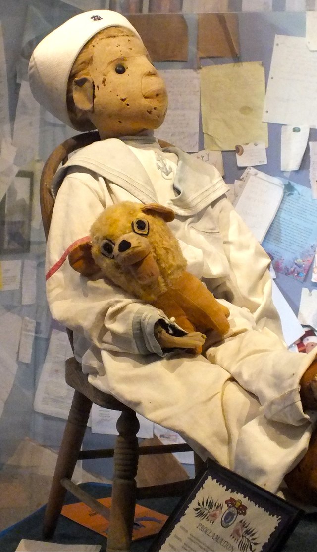 Robert the Doll, a large felt doll in a white sailor suit holding a small stuffed toy, seated in a glass display case