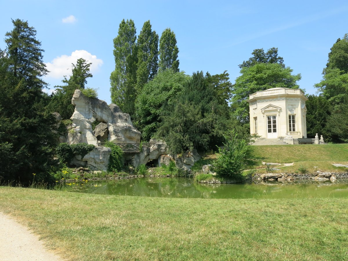 A small white octagonal pavilion beside a pond and an artificial rock formation in a park