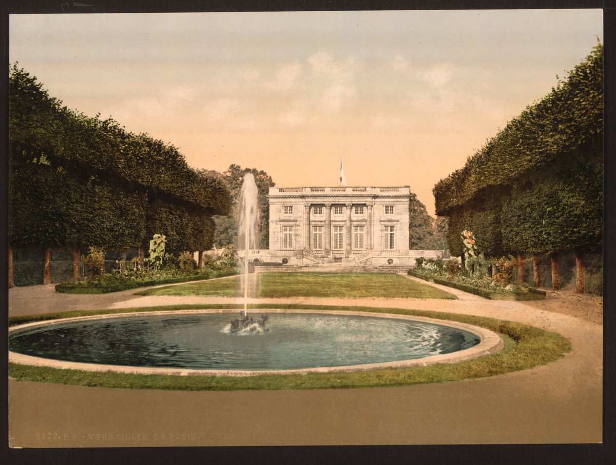 A color photochrom of the Petit Trianon at Versailles seen across a round fountain pool, framed by clipped hedges